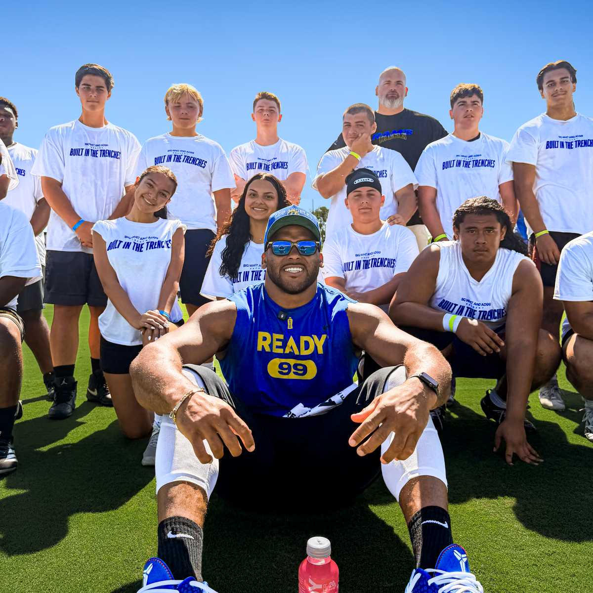 A group of athletes posing outdoors on a sunny day, with the central figure wearing a blue 'READY 99' jersey and blue Nike sneakers, surrounded by teammates in white 'built in the trenches' t-shirts. A pink grapefruit clear protein water bottle is placed in front of him on the green turf under a clear blue sky.